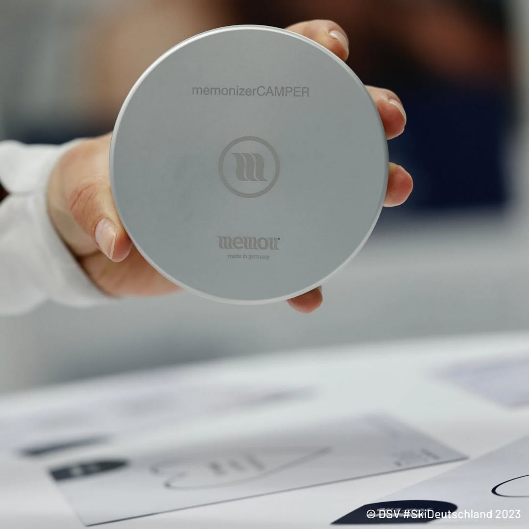 Hand houdt een ronde, zilverkleurige memonizerCAMPER vast, met memon-logo en de tekst “made in germany”.