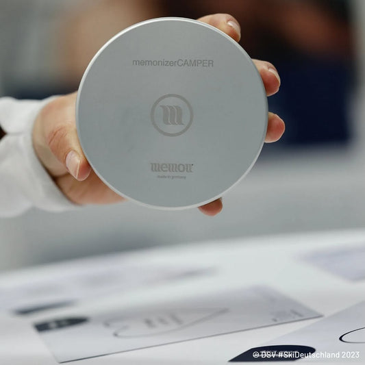 Hand houdt een ronde, zilverkleurige memonizerCAMPER vast, met memon-logo en de tekst “made in germany”.