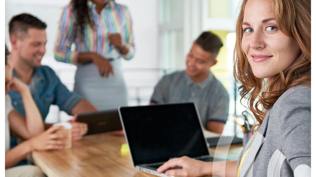 Vrouw achter een laptop kijkt lachend in de camera; op de achtergrond overlegt een team aan een kantoortafel.