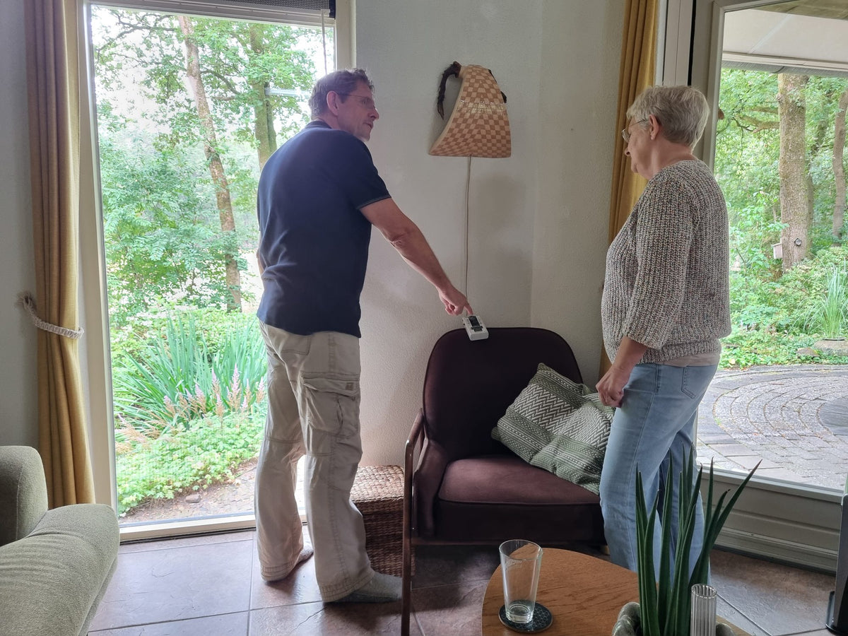 Woonbioloog Erik Leemkuil meet in een woonkamer met een handmeter, terwijl een klant toekijkt.